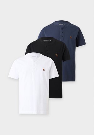 Mand med flettet hår iført runde solbriller, sort henley-shirt med korte ærmer, lyse revnede vidde-ben jeans og hvide sneakers.