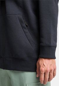 Sudadera con cremallera de un azul marino oscuro, con puños y dobladillo acanalados, que cuenta con un bolsillo lateral y una tela texturada. Mano visible en la esquina inferior derecha.