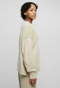 Beige oversized sweater with ribbed cuffs and hem, featuring a textured knit pattern. Long sleeves and relaxed fit.