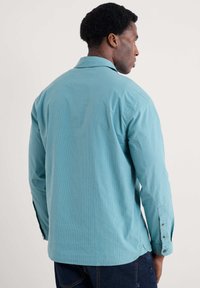 Koszula zapinana na guziki w jasnym odcieniu turkusowym z pionowymi paskami, z długimi rękawami zakończonymi mankietami na guziki, klasycznym kołnierzykiem i lekko zaokrąglonym dołem.