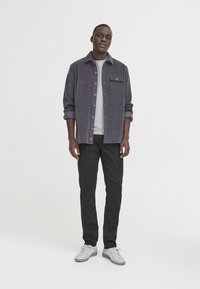 Giacca camicia da uomo in velluto a coste grigia, abbottonata, con taschino sul petto. Indossata sopra una maglietta grigio chiaro, abbinata a pantaloni neri e sneakers grigie.