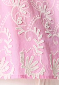 Tissu rose avec une broderie florale blanche complexe et des motifs texturés, mettant en valeur des points détaillés et des motifs de feuilles.