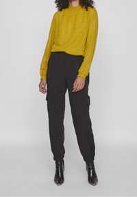 Blouse jaune à motif en chevrons avec un col montant et des poignets froncés, associée à un pantalon cargo noir et fuselé avec des poches latérales. Bottines noires.