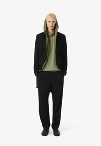 Blazer nero con una giacca verde con cappuccio sotto, indossato sopra una polo verde, abbinato a pantaloni neri e sneaker nere.