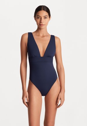 Maillot de bain une pièce bleu marine avec un décolleté en V profond, des larges bretelles et un design sans coutures. Fabriqué en matériau lisse ; des ouvertures de jambe à haute coupe mettent en valeur la silhouette.