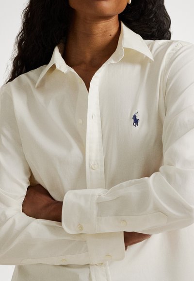 Kvinne med sammenkryssede armer iført en off-white skjorte med knapper og en liten mørkeblå polo-spillerlogo på brystet.