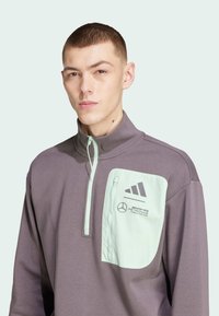 Felpa grigia con zip, dotata di una tasca verde menta, logo Adidas e branding Mercedes sul petto. Tessuto morbido con colletto alto.