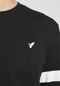 Svart bomull T-shirt med en vit broderad fågel-logotyp på vänster bröst och ett vitt horisontellt rand över den nedre ärmen.