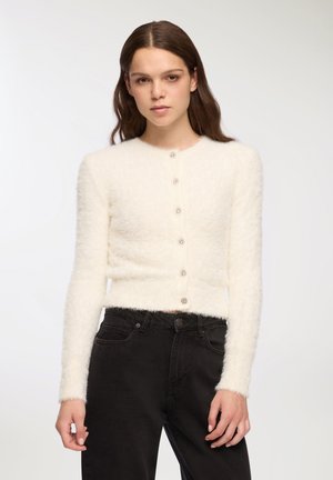 Cardigan bianco e peloso con collo rotondo, maniche lunghe e cinque bottoni decorativi sul davanti. Indossato con jeans neri a vita alta.