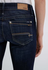 Jeans di denim scuro con vita media, dotati di una toppa in pelle marrone con il logo "mavi", cuciture gialle e due tasche posteriori con accenti metallici.