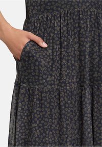 Dunkles Textilkleid mit gestuftem Design und gepunktetem Muster in hellbeige sowie seitlichen Taschen, die in die Naht integriert sind.