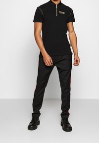 Zwarte poloshirt met een gouden voorrits en logo, gecombineerd met zwarte broeken met rode zijstreep. Zwarte veterschoenen maken de outfit compleet.