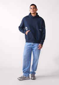 Navy blue hoodie with a front pouch pocket and graphic accents, paired with light blue straight-leg jeans and gray athletic shoes.