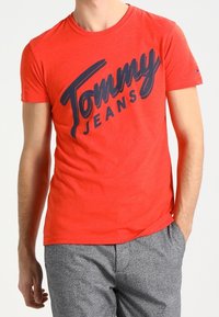 Czerwona bawełniana koszulka z krótkim rękawem, z dużym granatowym napisem "Tommy Jeans" w kursywie, okrągłym dekoltem.