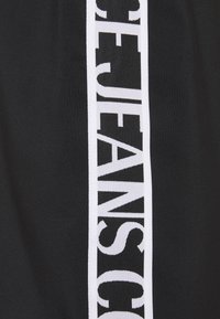 Svart tyg med ett vertikalt vitt rand som innehåller stor, fet svart text som säger "JEANS" delvis synlig.