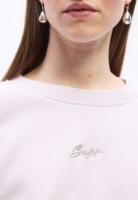 Γυναίκα με ίσια καστανά μαλλιά, φοράει ασημένια σκουλαρίκια σε σχήμα σταγόνας και ανοιχτό ροζ τοπ με κέντημα τη λέξη "Boss" στο στήθος.