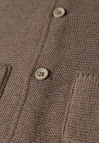 Brauner Strickstoff mit strukturierter Oberfläche, mit einer Frontknopfleiste aus einer cremefarbenen Taste und einer kleinen Tasche.