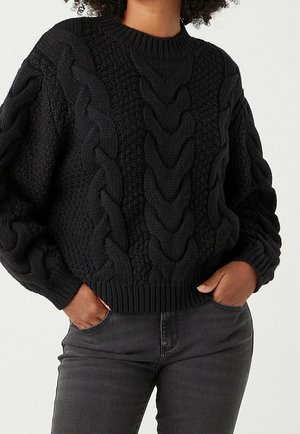 Jersey negro grueso de punto con grandes patrones trenzados de cable, usado con vaqueros gris oscuro, manos en los bolsillos, sobre fondo claro y liso.