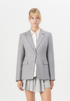 Gray tailored blazer with notch lapels, single-button closure, and two front pockets, worn over a light-colored shirt and pleated skirt.