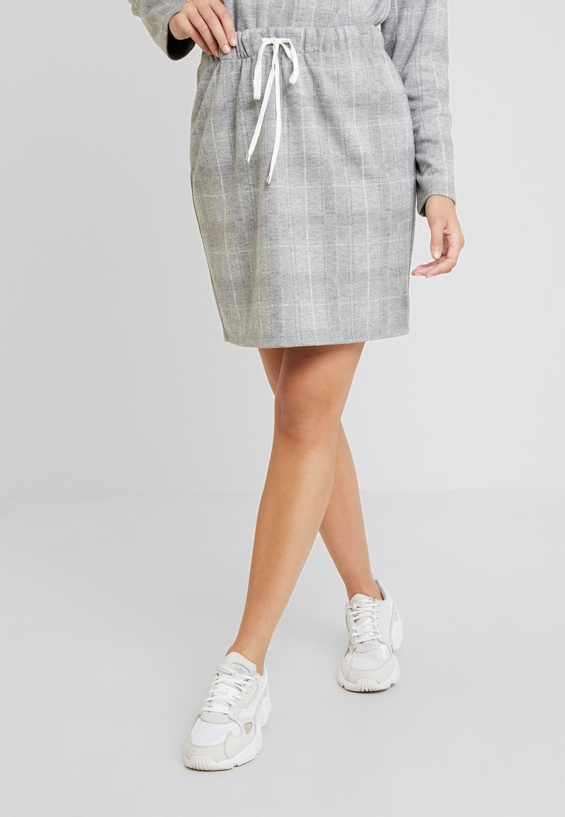 Femme portant une jupe grise à carreaux avec un cordon blanc et des baskets blanches, debout avec une jambe légèrement croisée devant l'autre.