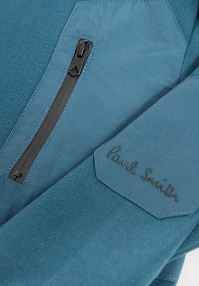 Giacca in pile blu con una tasca con zip nera, tessuto testurizzato e il logo "Paul Smith" in rilievo su un'apposita patch in contrasto.