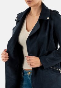 Giacca in tessuto simile alla suede di colore blu navy, con ampio colletto, hardware argentato e tasca laterale con zip, indossata aperta sopra una maglietta a costine chiara.