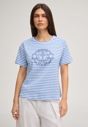 Frau trägt ein hellblau-weiß gestreiftes T-Shirt mit einem blauen dekorativen Emblem und dem Schriftzug "JOOP!", kombiniert mit weißen Hosen.