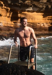 Een bloot bovenlijf man staat op een metalen ladder bij het water, gekleed in zwarte zwembroek met witte patronen, tegen een rotsachtige oranje achtergrond.
