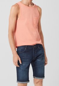 Top sin mangas de coral claro con un bolsillo en el pecho, combinado con jeans de denim oscuro. Los jeans tienen un dobladillo enrollado y un diseño clásico de cinco bolsillos.
