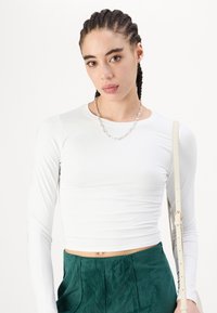Fehér, hosszú ujjú, crop top sima anyagból, zöld corduroy, bőszárú nadrággal párosítva. Figyelemre méltó ezüst lánc nyaklánc és világos táska.