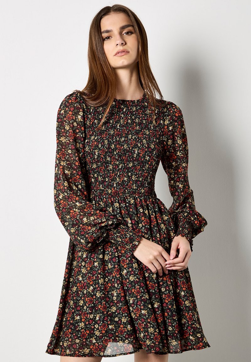 Czarna sukienka w kwiaty z marszczonym przodem, długimi bufkami i rozkloszowanym dołem, zdobiona czerwonymi, zielonymi i żółtymi wzorami kwiatowymi.
