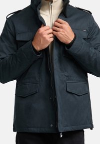 Mørkeblå frakke med tekstureret overflade, der har frontzip og knaplukning, to sidelommer og skulderstropper.