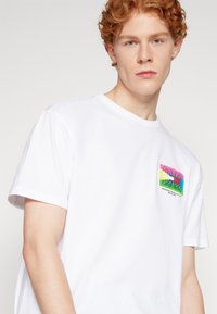 Camiseta blanca de algodón con un estampado gráfico colorido en el lado izquierdo del pecho que presenta bandas de color rosa, amarillo, verde y azul con detalles de texto.