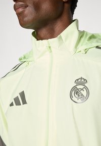 Homme portant une veste Real Madrid vert clair avec col zippé et logo Adidas sur l'épaule et la poitrine.