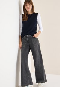 Granatowy top bez rękawów noszony na białej koszuli z długim rękawem, zestawiony z szerokimi spodniami z szarego denimu z bocznymi kieszeniami i wyblakłym wykończeniem.