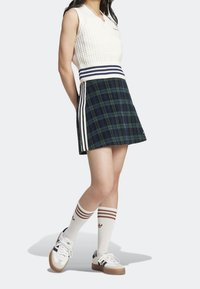 Bílé pletené tílko bez rukávů s výstřihem do V, doplněné tmavou kostkovanou sukní s bílými bočními pruhy a hnědými ponožkami s pruhy jako akcentem.