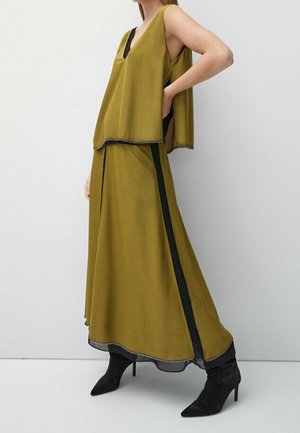 Femme portant un haut sans manches vert olive et une longue jupe assortie avec des bandes latérales noires et des bottines noires à talons.