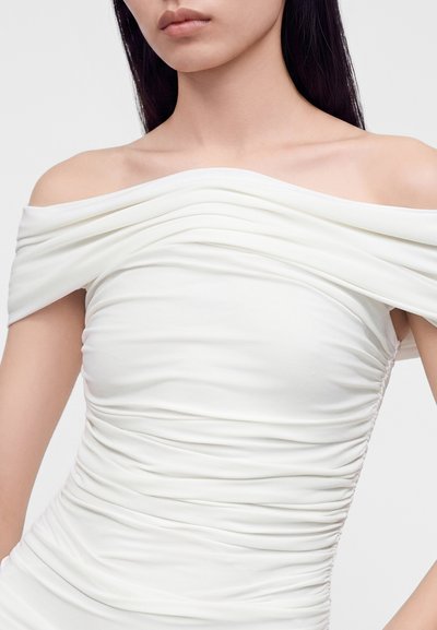 Kvinde iført hvid off-shoulder kjole med rynker, som viser bare skuldre og øvre bryst, med lige mørkt hår.