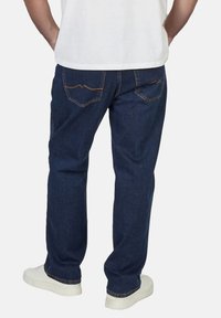 Hombre vestido con jeans de mezclilla azul oscuro, zapatillas blancas y una camiseta blanca, de pie con las manos en los bolsillos traseros frente a un fondo liso.