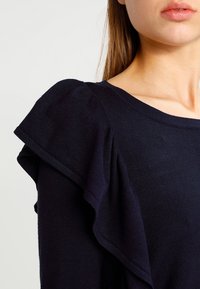 Top lavorato a maglia blu navy con maniche a volant e scollatura rotonda, che mette in risalto una trama liscia e un design strutturato.