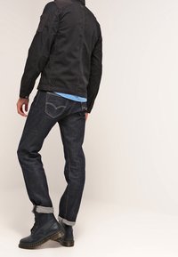 Giacca nera casual con toppe sulle spalle, maglione blu sotto, jeans di denim scuri e stivali neri stringati con suola in gomma.