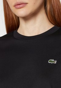 Schwarzes Baumwoll-T-Shirt mit Rundhalsausschnitt, das auf der linken Brustseite mit einem kleinen grünen und weißen Krokodil-Logo bestickt ist. Glatte Textur.
