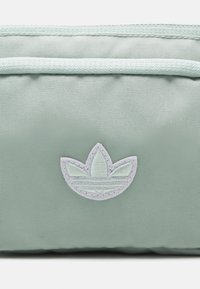 Mátově zelená taštička vyrobená z texturovaného nylonu, s bílým vyšitým logem. Design zahrnuje zipový uzávěr a jemné vodorovné pruhy.