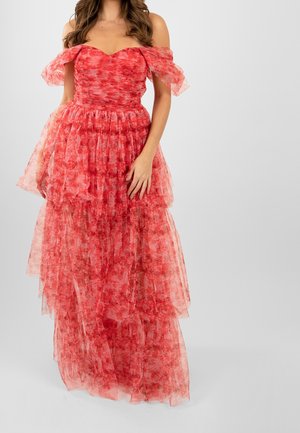 Woman wearing off-shoulder, layered red floral tulle gown with ruffled sleeves and fitted bodice, standing against a plain background.