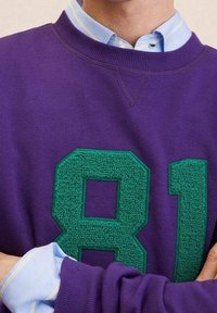 Sudadera morada con un gran "81" texturizado en verde en la parte delantera, llevada sobre una camisa de color azul claro con cuello y rayas blancas y azules.