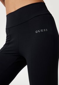 Melnas leggos, kas izgatavoti no gluda, elastīga auduma, ar platām jostām un sudraba "GUESS" logotipu kreisajā augšstilbā.