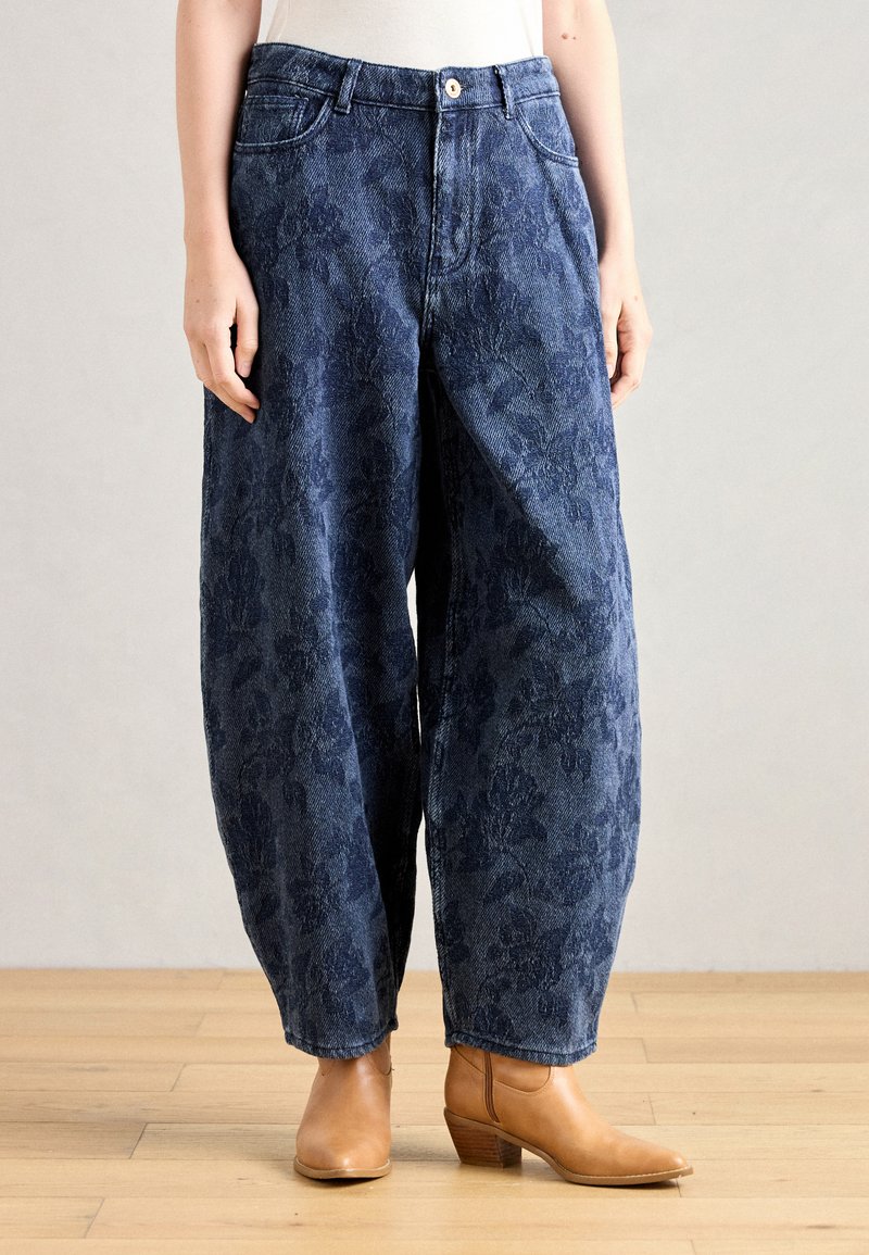 Széles lábú kék farmernadrág, texturált virágmintával, magas derékkal és elülső gombos záródással. Barna bokacsizmákkal párosítva.