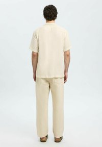 Camicia beige a maniche corte con taglio dritto, abbinata a pantaloni beige larghi e sandali, vista da dietro.