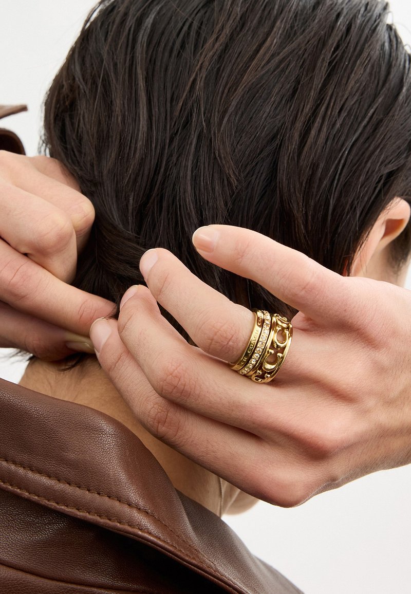 Main avec trois bagues en or ajuste des cheveux foncés, personne portant une veste en cuir marron, gros plan sur les mains et les cheveux.