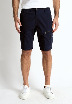 Mand iført sorte cargoshorts med lynlås- og knaplommer, hvide sneakers og en hvid skjorte, stående på en hvid baggrund.